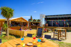 Holzterrasse mit Tisch und Getränken, im Hintergrund Campingplatz mit überdachtem Bereich, Spielgeräten und Sonnenschirmen.
