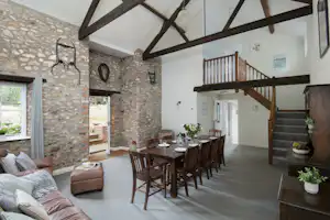 Wohnbereich mit Esstisch und Stühlen, Steinwand und Sofa, Holztreppe und offene Deckenbalken im Ferienhaus.