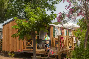Holz-Bungalow mit Terrasse im Garten, Stühle und Tisch vor dem Eingang, Bäume und blühende Sträucher im Vordergrund.