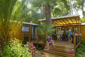 Holz-Blockhaus mit Terrasse im Campinggarten, umgeben von Palmen und Grünpflanzen, mit Kindern auf der Veranda.