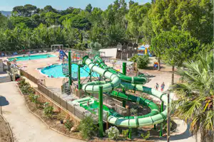 Großes Camping-Schwimmbecken mit grünen Wasserrutschen, Liegen am Rand und Bäumen im Hintergrund.