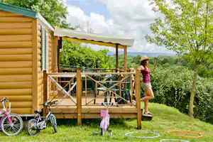 Gelber Wohnwagen mit Holzterrasse, davor Fahrrad und Spielzeug, im Grünen mit Bäumen und Blick ins Tal.