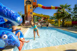 Familie im Camping-Pool mit blauen Wasserrutschen, Kinder planschen im Wasser vor gelben Rutschen und Liegestühlen.