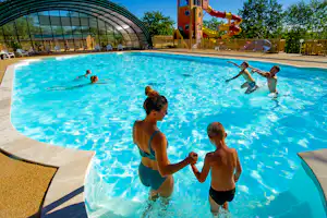 Kinder und Erwachsene im blauen Schwimmbecken eines Campingplatzes, im Hintergrund Spielbereich und Bäume.