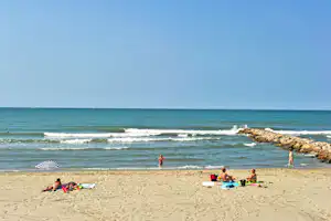 Sandstrand mit Badegästen, Stranddecken und Sonnenschirm, im Hintergrund Wellen und Felsen am Ufer.