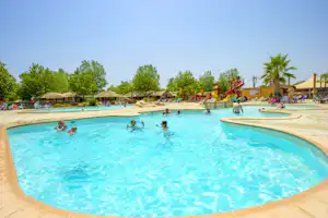 Großes Schwimmbecken auf dem Campingplatz mit Liegen, Sonnenschirmen und Palmen im Hintergrund.
