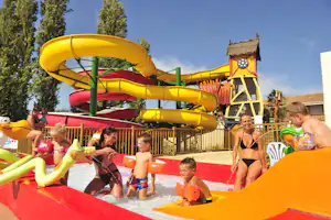 Kinder und Erwachsene auf einer großen gelben Wasserrutsche im Campingbereich, mit Holzplattform und Zaun, im Hintergrund Bäume.