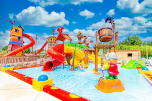 Wasserrutsche und Spielbereich im Campingpark mit bunten Rutschen, Planschbecken und Sonnenschirmen, daneben ein Holzhaus.