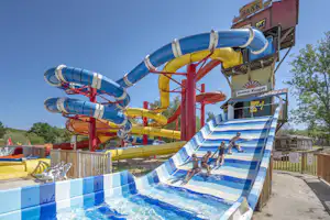 Wasserrutschen am Campingplatz: blaue und rote Rutschen über einem kleinen Becken, daneben ein Gebäude und Zaun.