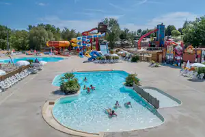 Großes Schwimmbecken mit Kindern im Wasser, daneben Sonnenliegen und ein Bereich mit roten Wasserrutschen.