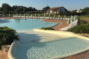 Camping-Pool mit türkisfarbenem Wasser, Sonnenliegen und Sonnenschirmen am Beckenrand, im Hintergrund Bungalows und Bäume.