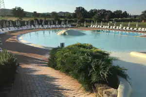 Schwimmbecken auf einem Campingplatz mit gepflastertem Weg, Liegen am Beckenrand und Bäumen im Hintergrund.