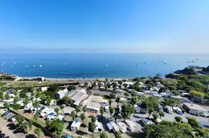 Campingplatz mit Stellplätzen und Wohnwagen, Blick auf das Meer und Boote, umgeben von Bäumen und Wegen.