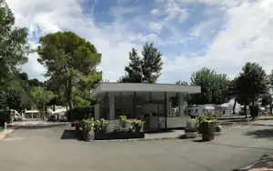 Rezeption mit überdachtem Eingang auf dem Campingplatz, gepflasterter Platz, Bäume und Pflanzenkübel im Vordergrund.