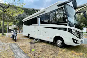 Weißes Wohnmobil auf einem Campingplatz-Stellplatz, daneben Motorrad, gepflasterter Weg und Bäume im Hintergrund.