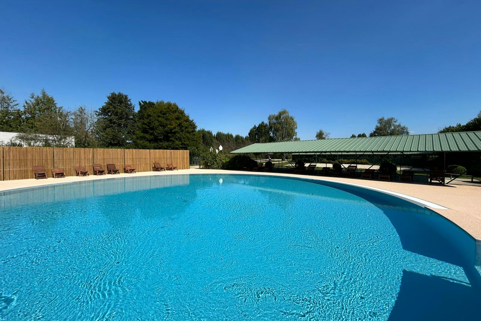 Blauer Campingpool mit Liegen unter grünem Dach, Holzsichtschutzwand und Bäumen im Hintergrund.