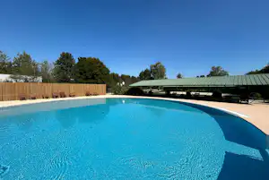Blauer Campingpool mit Liegen unter grünem Dach, Holzsichtschutzwand und Bäumen im Hintergrund.
