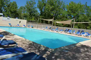 Blauer Pool mit Liegestühlen und Sonnensegeln am Campingplatz, dahinter Holzsichtschutz und Bäume im Hintergrund.