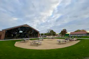Holzgebäude mit großer Glasfront, Terrasse mit Tischen und Stühlen, Rasenfläche und weitere Gebäude auf dem Campingplatz.