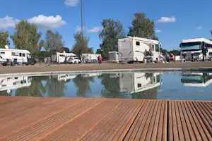 Wohnmobile auf dem Campingplatz neben einem kleinen Becken, mit Spiegelung im Wasser und Bäumen im Hintergrund.