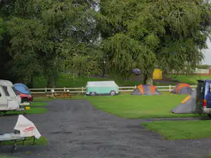 Campingplatz mit mehreren Zelten und einem Wohnwagen auf einer grünen Wiese, umgeben von Bäumen und einem Weg.