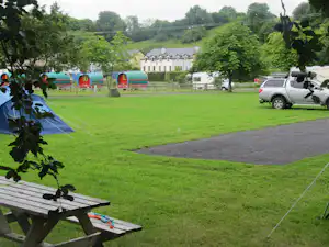 Grüner Campingplatz mit Rasen, Picknicktisch und Stellplatz für Wohnwagen, daneben ein Auto und Bäume im Hintergrund.