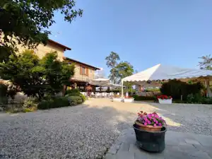 Terrasse am Campingplatz mit Zeltüberdachung, Kiesweg, Blumenkübel und Wohngebäude mit Fenstern und Dach.