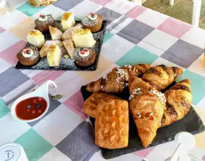 Frühstückstisch mit Croissants und Gebäck auf einer karierten Decke, daneben Schälchen mit Sauce und kleine Kuchenstücke.