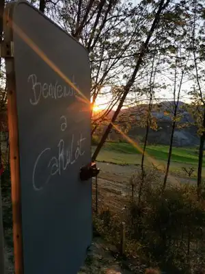 Schild mit handschriftlicher Aufschrift am Campingplatz, im Hintergrund Wiese, Bäume und untergehende Sonne.