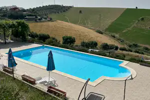 Blauer Campingpool mit Liegen und Sonnenschirmen, daneben gepflasterte Terrasse und Blick auf grüne Hügel und Felder.