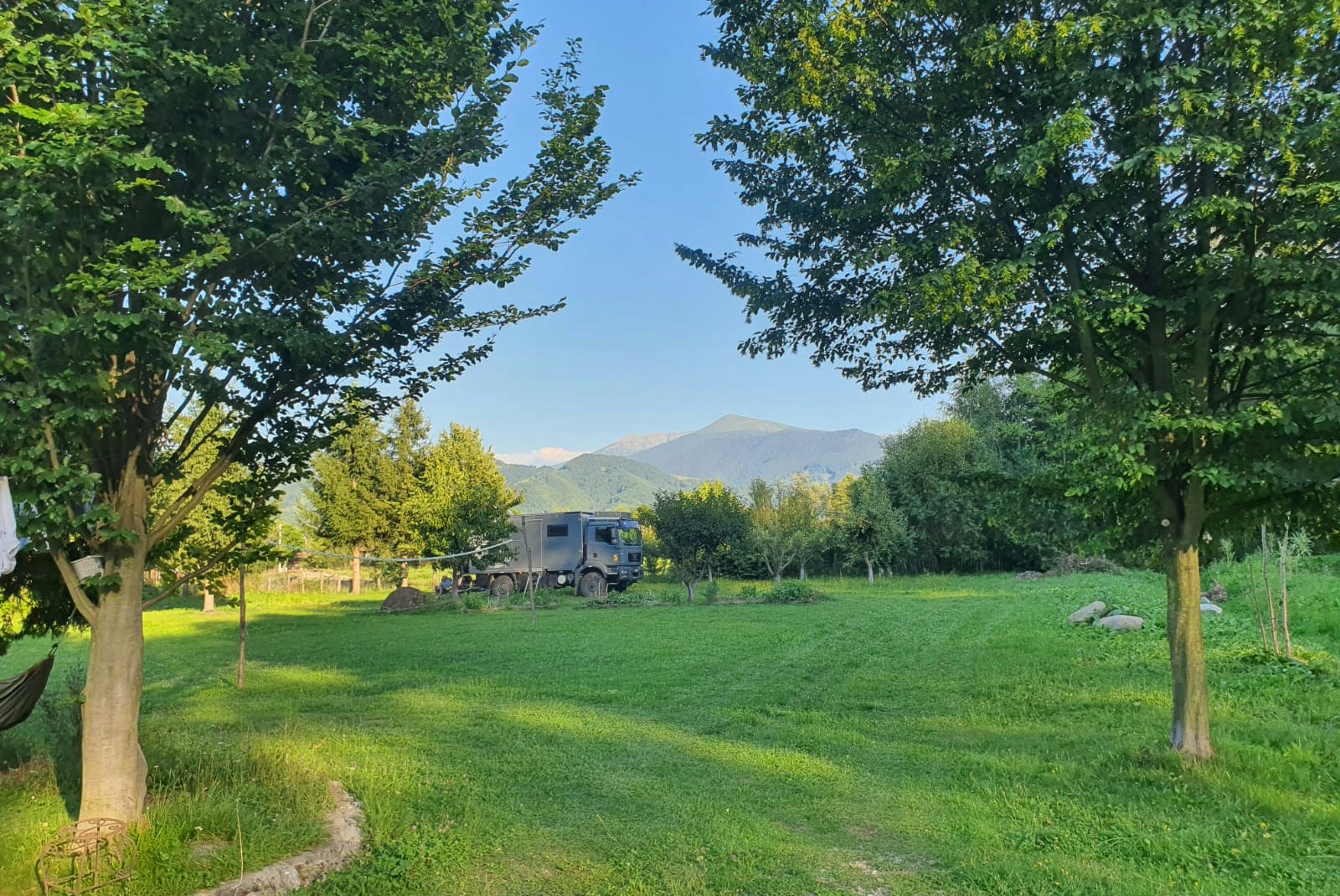 Grüne Campingwiese mit Bäumen, Weg und einem Wohnwagen im Hintergrund, dahinter Berge in der Ferne.
