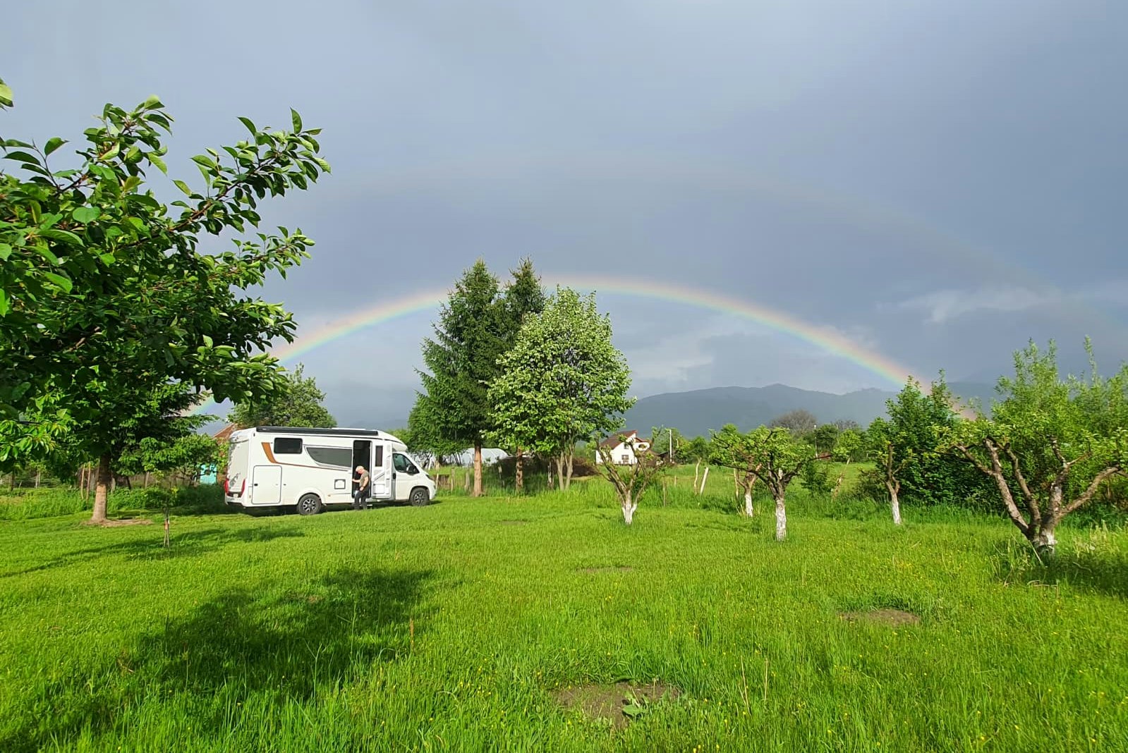 Wohnmobil auf einer grünen Campingwiese, Obstbäume und ein Regenbogen am Himmel vor bewaldeten Hügeln.