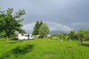 Wohnmobil auf einer grünen Campingwiese, Obstbäume und ein Regenbogen am Himmel vor bewaldeten Hügeln.