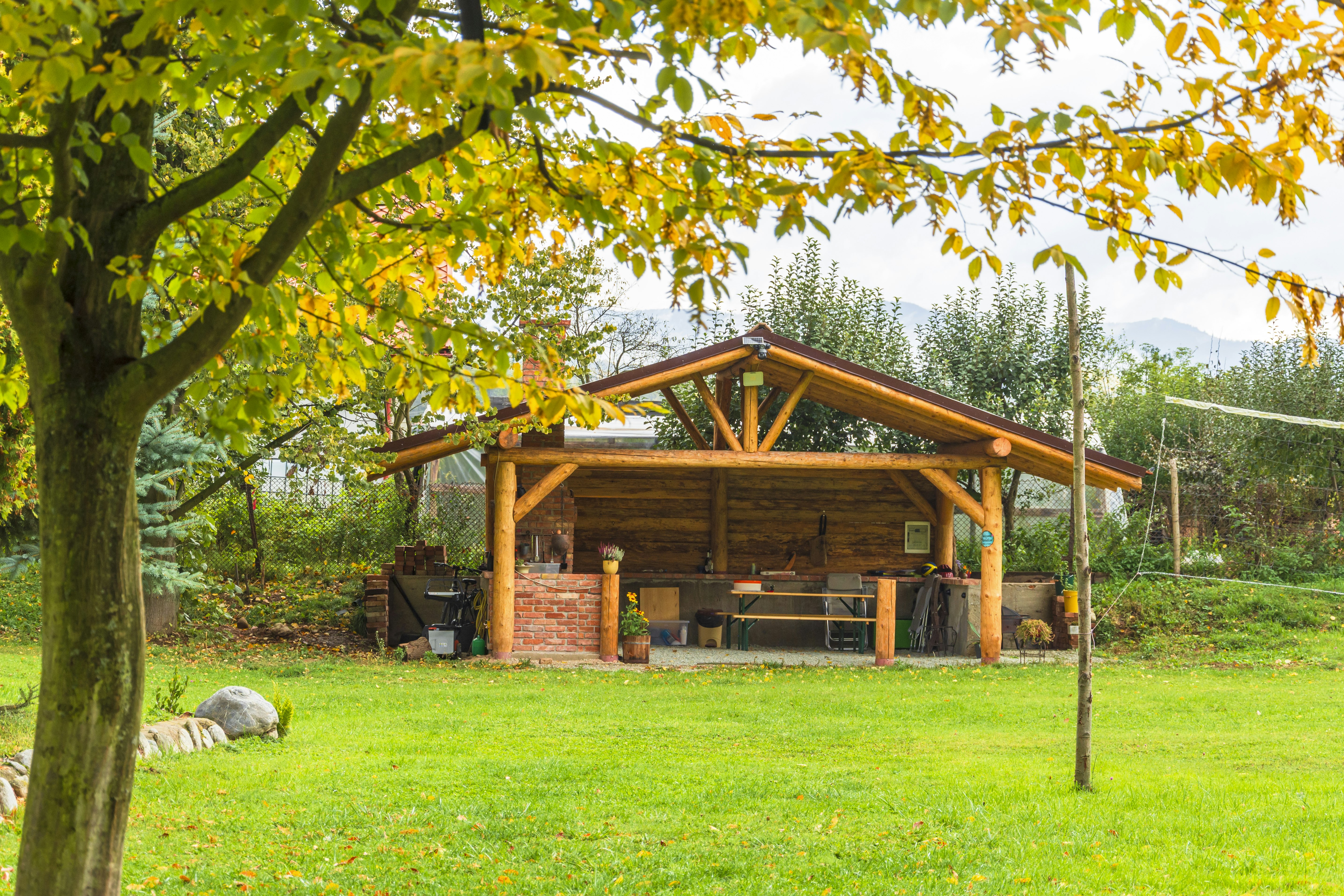 Holzunterstand mit überdachter Sitzbank und Grill auf einer grünen Wiese, im Hintergrund Bäume und ein See.