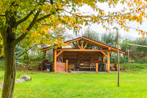 Holzunterstand mit überdachter Sitzbank und Grill auf einer grünen Wiese, im Hintergrund Bäume und ein See.