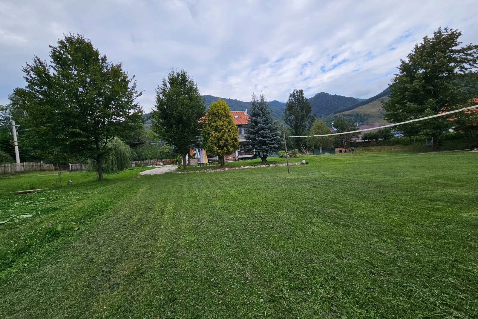 Weite Campingwiese mit Kiesweg, Bäumen und Blick auf Berge, im Hintergrund Gebäude und Zelte.
