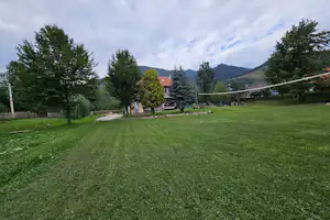 Weite Campingwiese mit Kiesweg, Bäumen und Blick auf Berge, im Hintergrund Gebäude und Zelte.