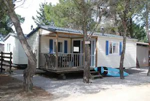 Mobilheim mit Holzterrasse, weißen Wänden und Fenstern, umgeben von Bäumen und Kiesweg am Campingplatz.