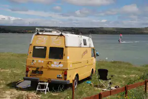 Gelbes Wohnmobil am Seeufer mit Markise, Campingstuhl und Tisch, im Hintergrund Windräder und bewachsene Hügel.