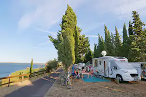 Wohnmobil auf dem Campingplatz am Meer, mit Vorzelt, Tisch und Stühlen, Fahrrad am Weg und Zypressen.