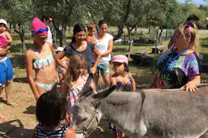 Familie streichelt einen Esel im Campingbereich, Kinder und Erwachsene am Zaun, im Hintergrund Bäume und ein Gehege.