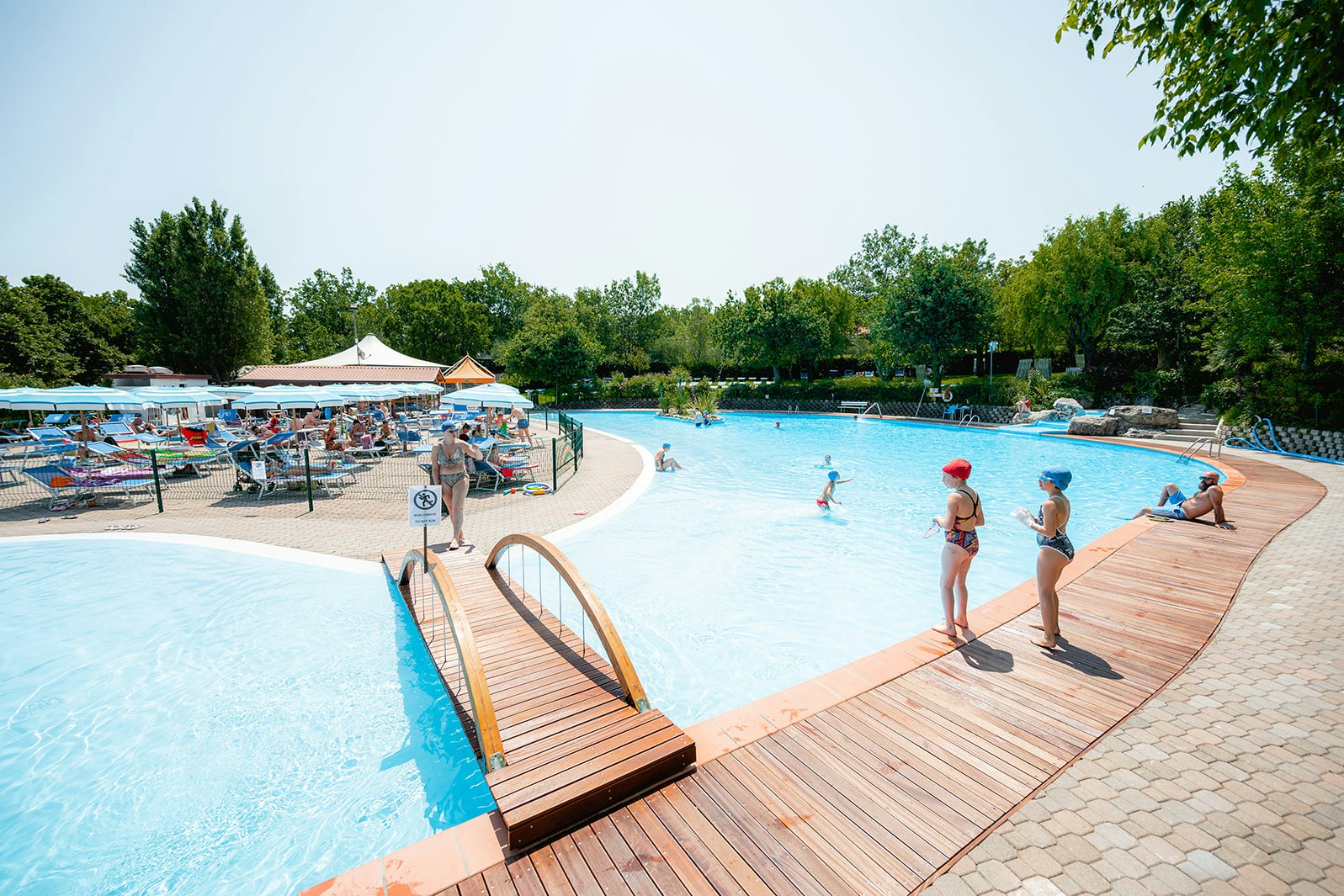 Camping-Pool mit Holzstegen, Liegen und Sonnenschirmen, im Hintergrund Bäume und ein ruhiger See.