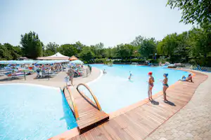 Camping-Pool mit Holzstegen, Liegen und Sonnenschirmen, im Hintergrund Bäume und ein ruhiger See.