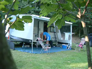 Wohnwagen auf dem Campingplatz im Grünen, mit Markise, Stühlen und Tisch vor dem Eingang, umgeben von Bäumen.