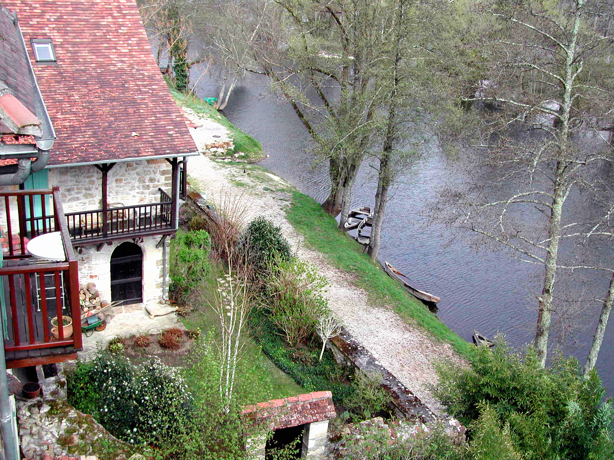 Campinghaus mit rotem Ziegeldach und Terrasse am Fluss, gepflasterter Weg und Bäume am Ufer.