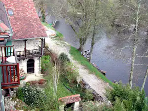 Campinghaus mit rotem Ziegeldach und Terrasse am Fluss, gepflasterter Weg und Bäume am Ufer.
