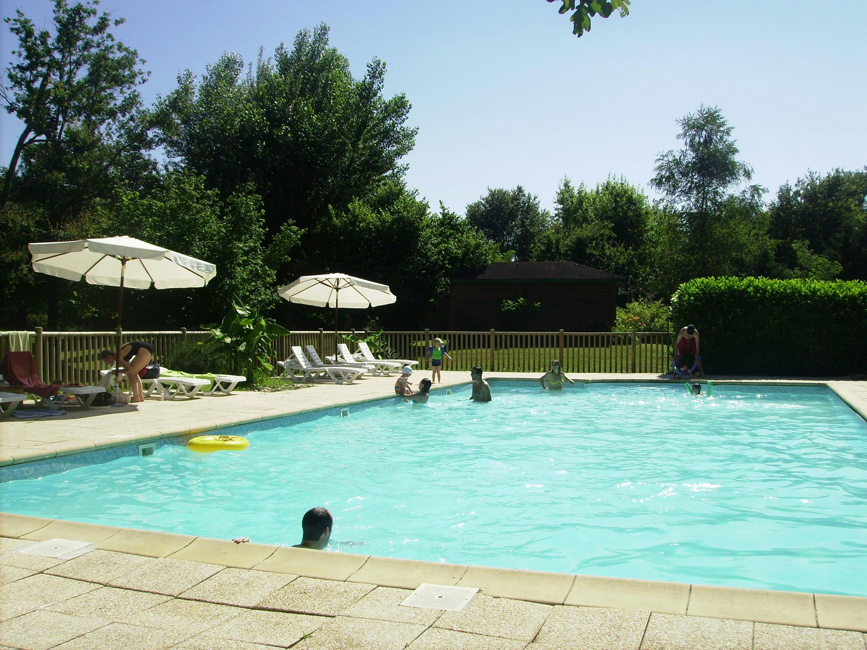 Camping-Pool mit türkisfarbenem Wasser, Sonnenliegen und Sonnenschirmen, umgeben von Hecken und Bäumen.