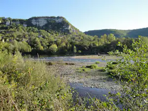 Ein Fluss mit Kiesufer und grünen Büschen, dahinter bewaldete Hügel und eine helle Felswand im Tal.