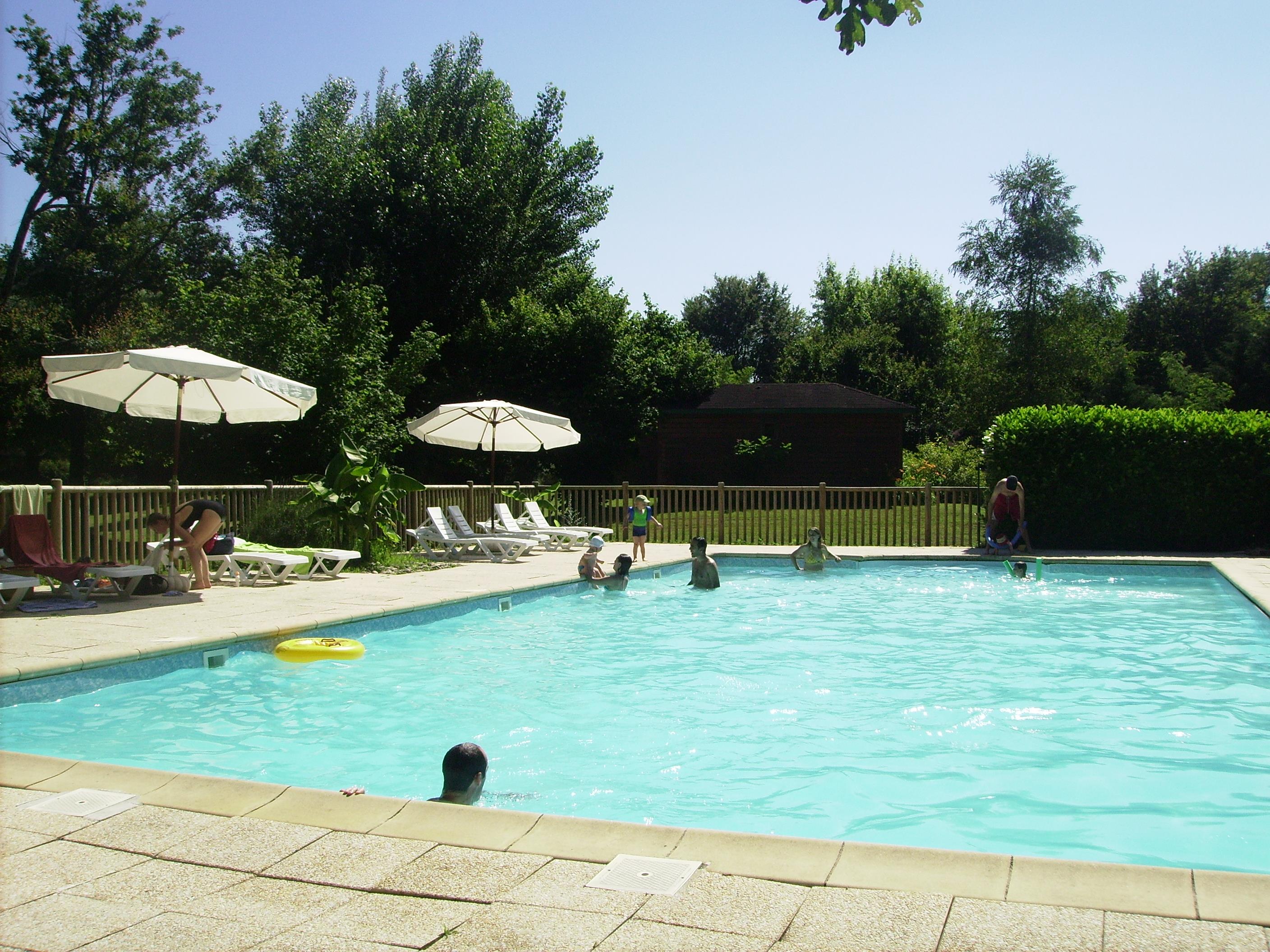 Campingpool mit türkisfarbenem Wasser, mehreren Liegen und Sonnenschirmen, im Hintergrund Hecken und Bäume.