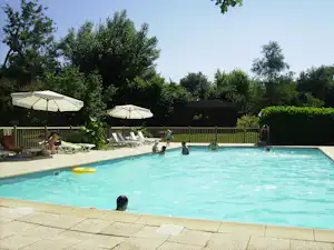 Campingpool mit türkisfarbenem Wasser, mehreren Liegen und Sonnenschirmen, im Hintergrund Hecken und Bäume.